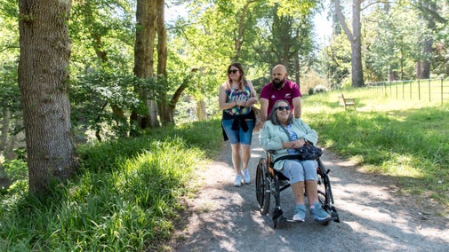 Three people, one a wheelchair user, explore a path through sunny woodland
